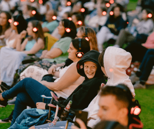A crowd of people sitting outdoors wearing headphones with red lights, attending what appears to be a silent event or concert.
