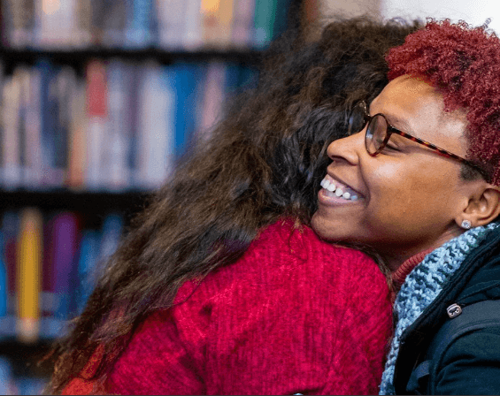 Two people warmly embracing in front of a blurred bookshelf, with one person smiling and wearing glasses and a red sweater.