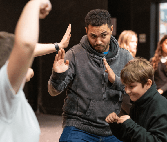 A man and two children practice playful martial arts poses in a classroom or studio, with others visible in the background.
