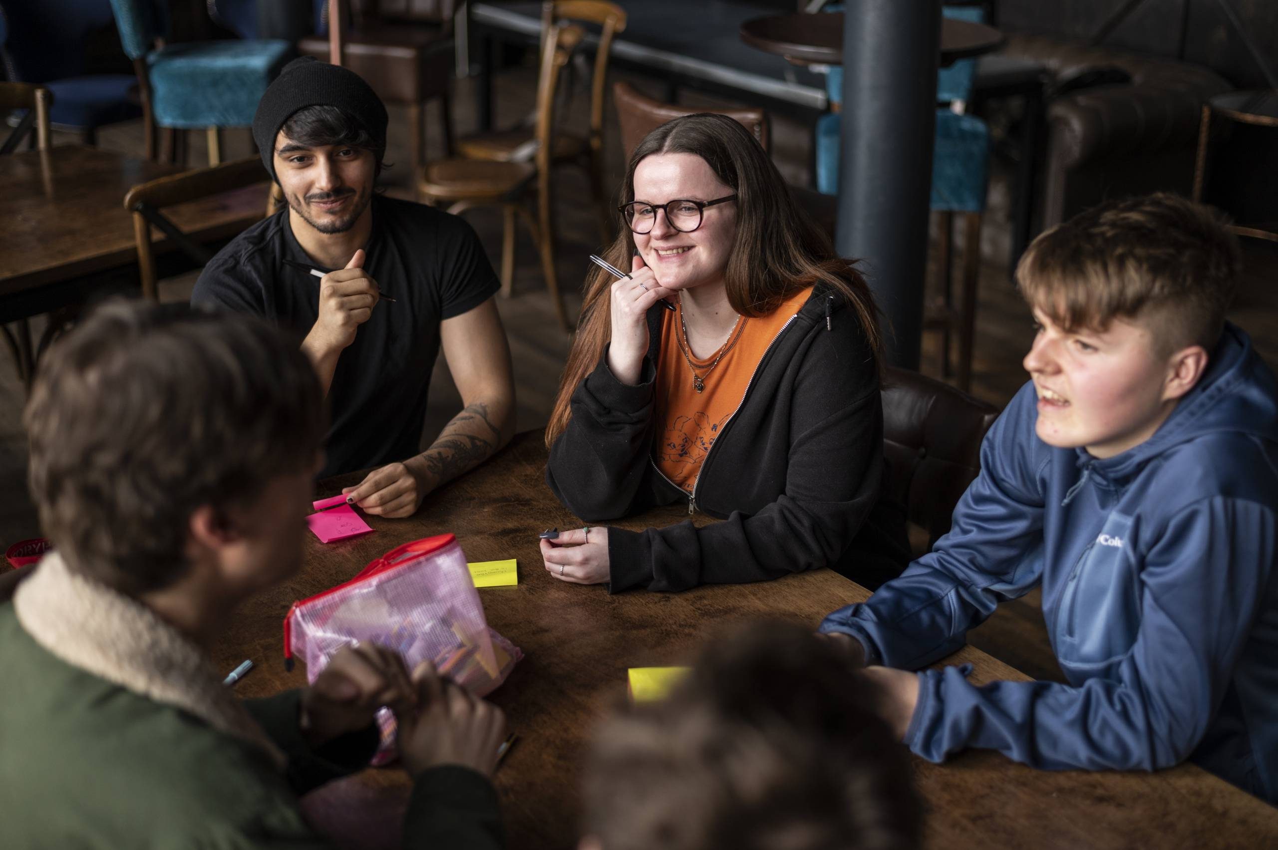 Four young people sit around a wooden table, talking and smiling, with colorful sticky notes and a pencil case in front of them.
