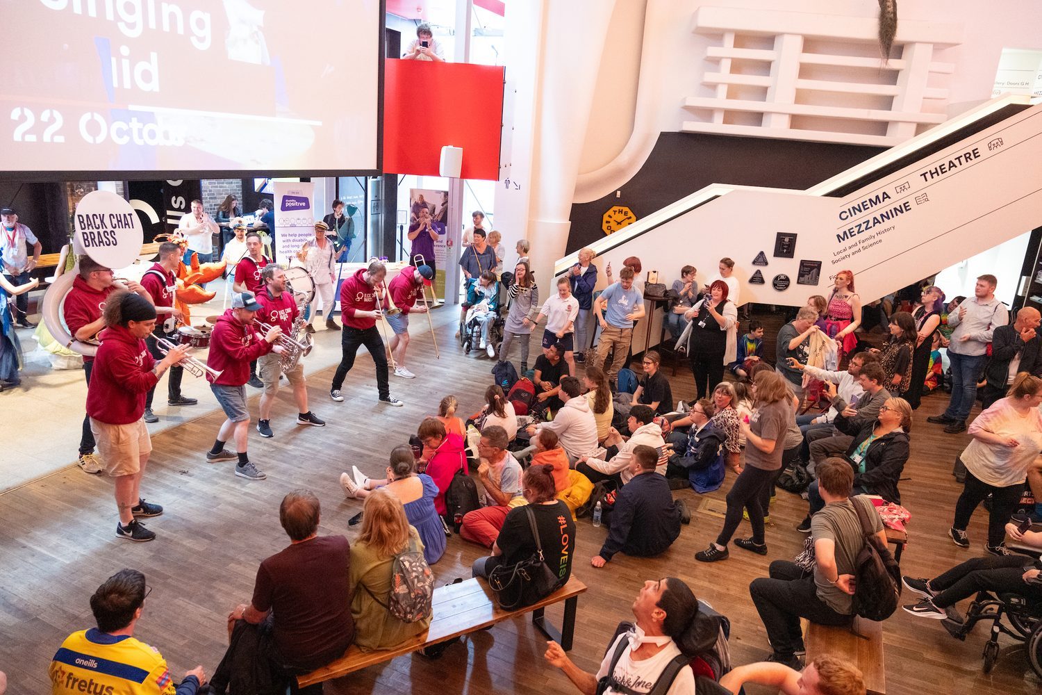 A band performs indoors for a seated and standing audience in a spacious venue with modern decor and a staircase in the background.